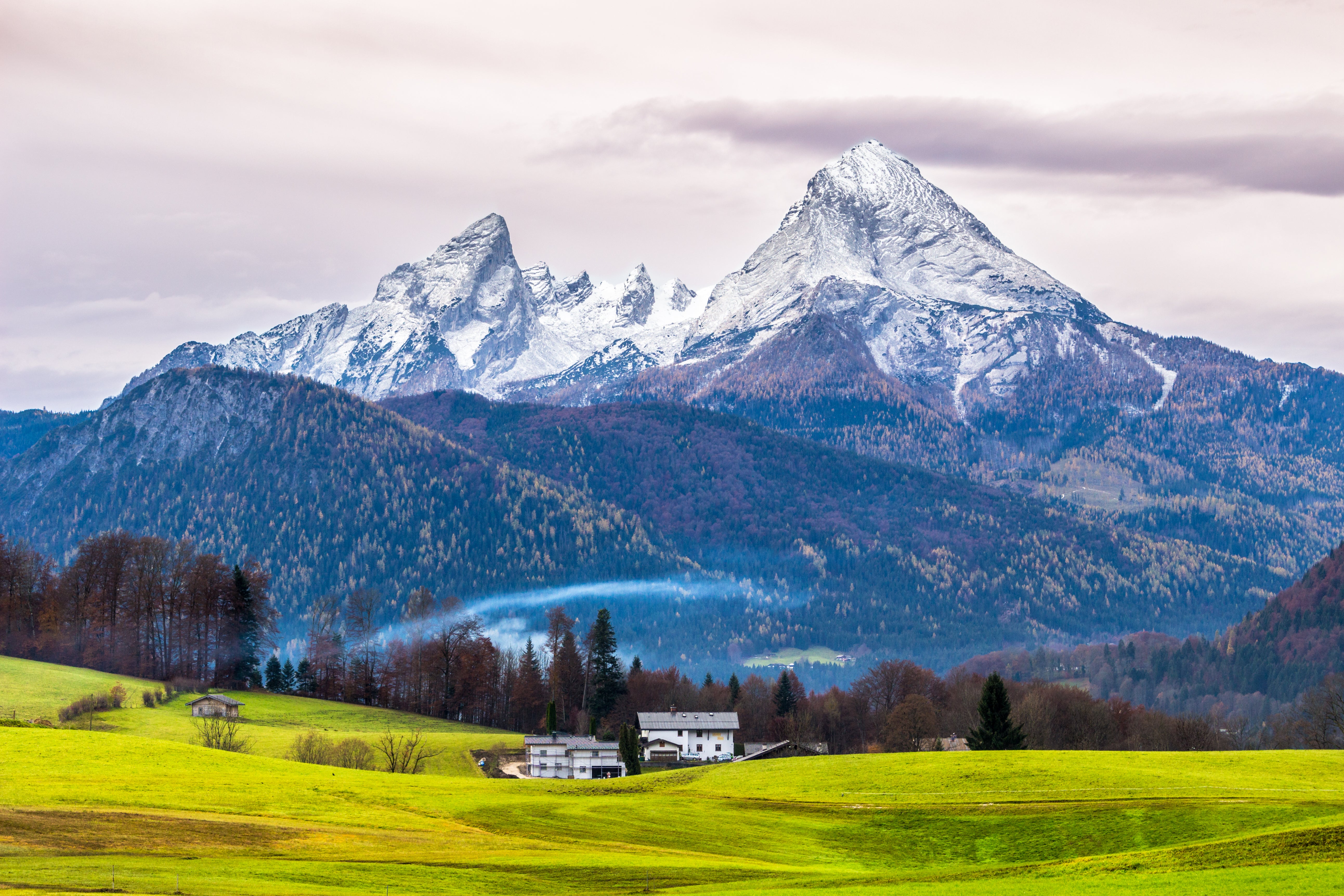 Verschneite Berggipfel im Hintergrund mit grünen Wiesen und Häusern im Vordergrund unter bewölktem Himmel.