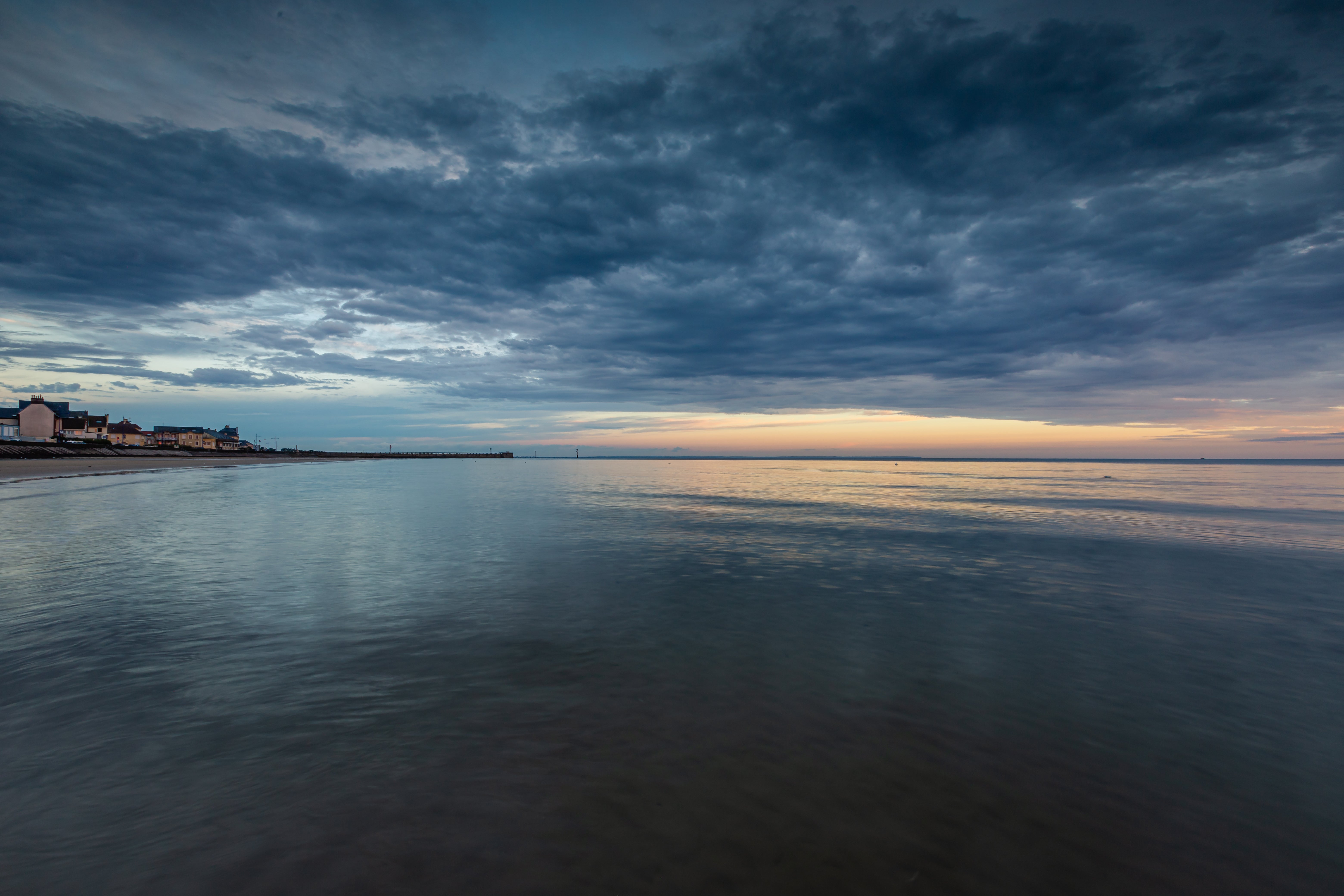 Dunkle Wolken über ruhiger See mit schwachem Sonnenlicht am Horizont und Häusern am linken Uferufer.