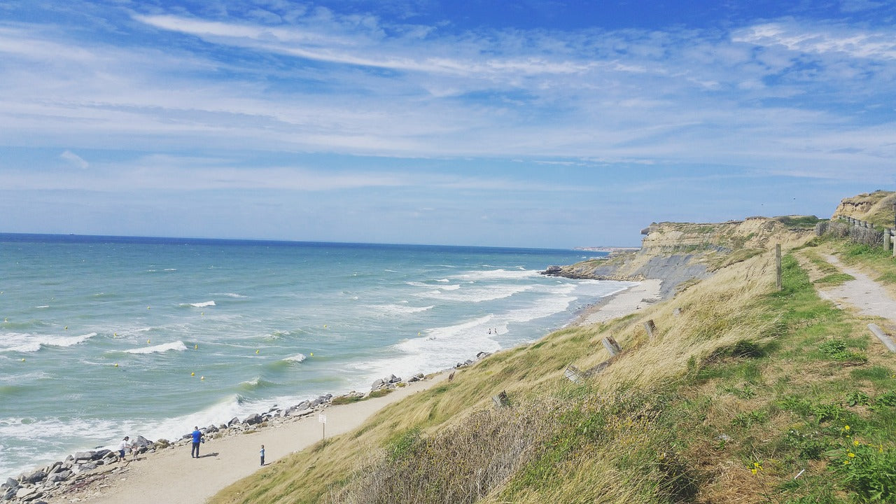 Küstenweg mit grasbewachsenen Klippen entlang eines ruhigen Strandes unter blauem Himmel mit vereinzelten Wolken