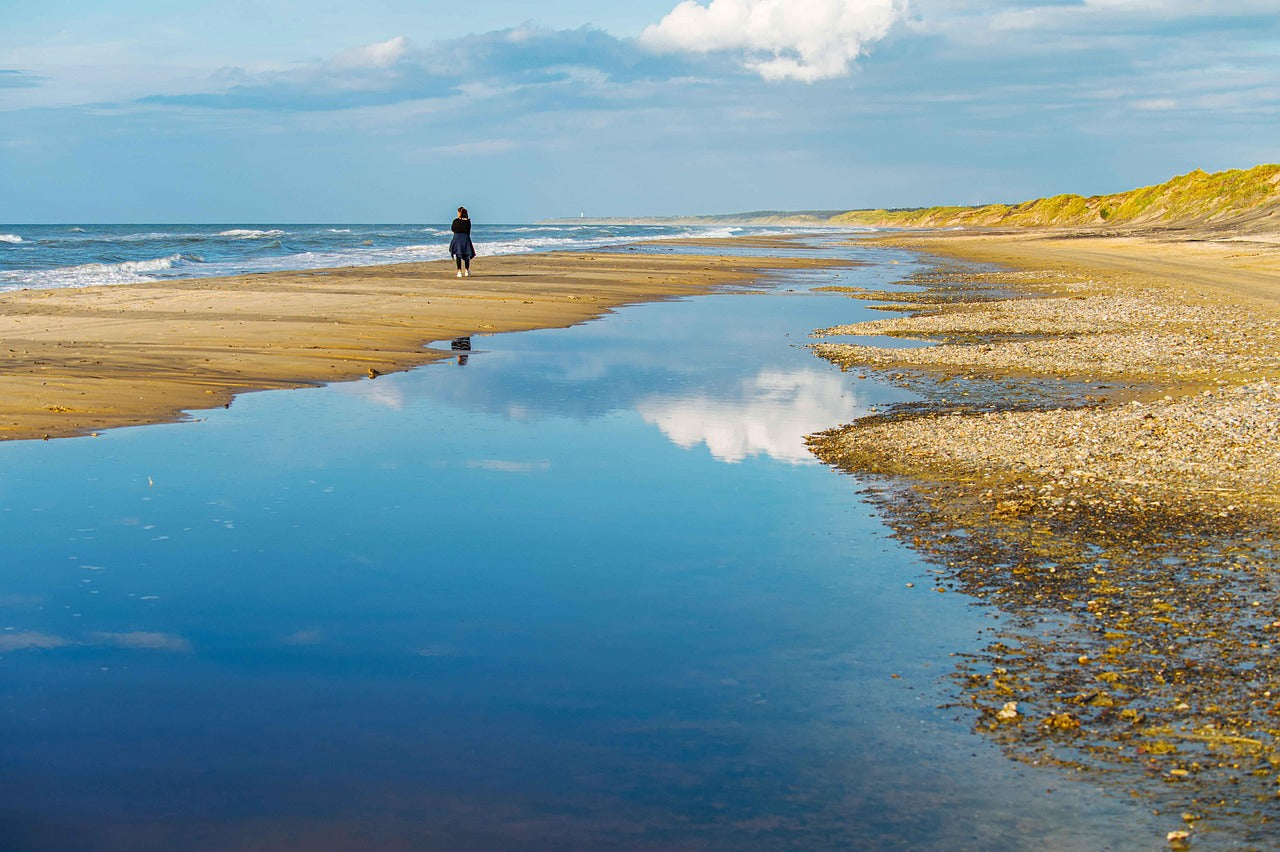 Weitläufiger Sandstrand mit Wasserpfützen, blauem Himmel und einer Person im Hintergrund beim Spaziergang.