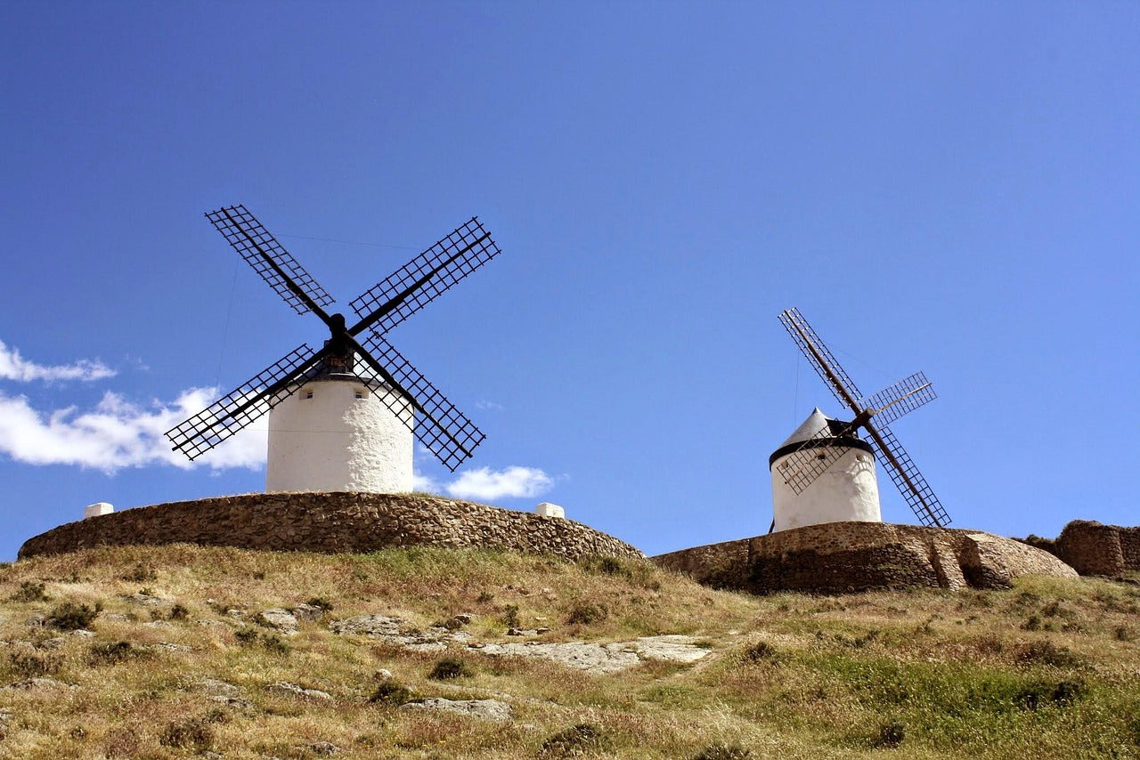 Zwei traditionelle Windmühlen auf einem hügeligen, grasbewachsenen Gelände unter klarem blauem Himmel.